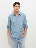 A person wearing the Mill Dress Shirt In End-on-End Stripe with rolled sleeves and light blue distressed jeans stands against a plain white background, hands in pockets, looking at the camera.