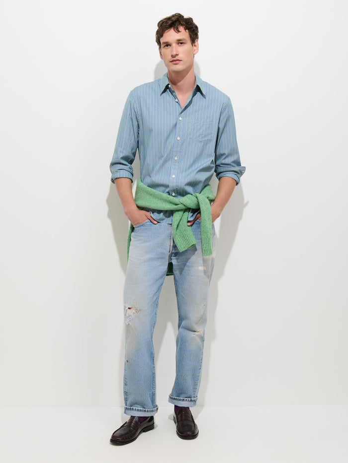 A person stands against a white background wearing the perfect shirt—the Mill Dress Shirt In End-on-End Stripe, a blue striped, button-down crafted from 100% cotton—paired with light blue ripped jeans, black shoes, and a green sweater tied around their waist.