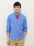 A young man with short dark hair stands against a white background, wearing the Mill Shirt in Cotton Poplin in blue over a red t-shirt and khaki pants, hands in his pockets, and a neutral expression.