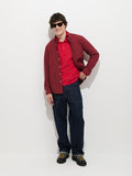 Wearing sunglasses, a person with short, dark hair stands and smiles against a plain white background, dressed in the Mill Shirt In Cotton Poplin (red) layered over a red polo, along with dark blue jeans and black shoes.