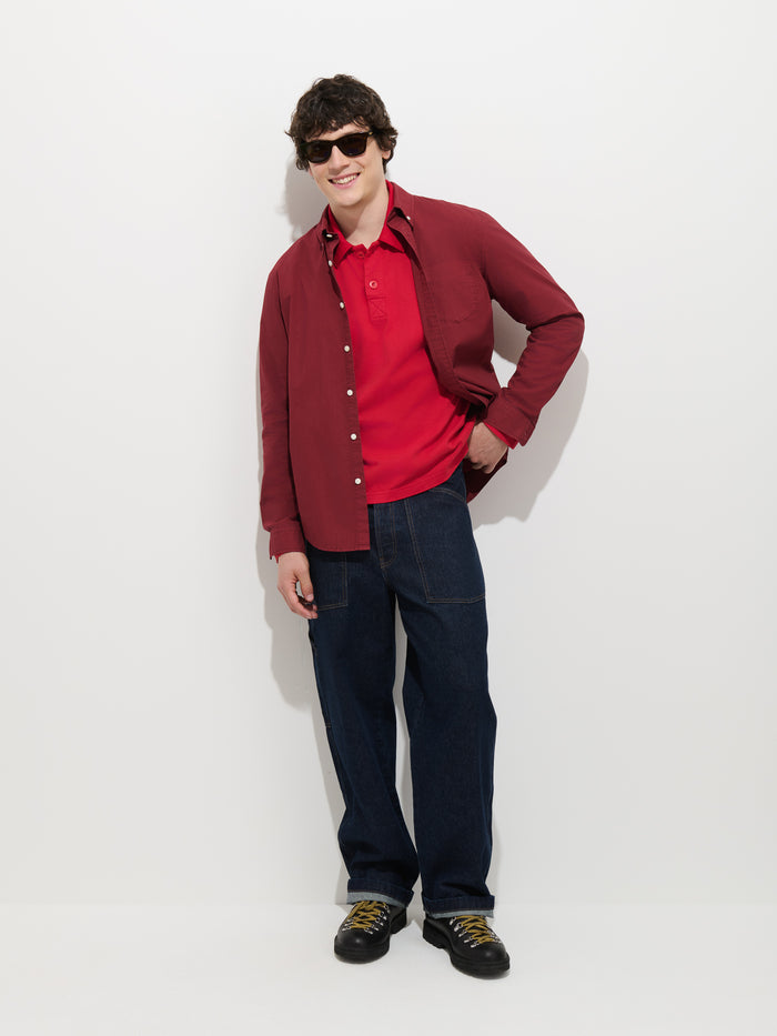 Wearing sunglasses, a person with short, dark hair stands and smiles against a plain white background, dressed in the Mill Shirt In Cotton Poplin (red) layered over a red polo, along with dark blue jeans and black shoes.