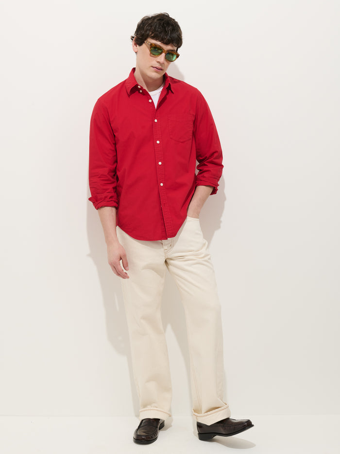 Wearing green sunglasses, the person stands with hands in pockets against a white background, dressed in the Mill Shirt In Cotton Poplin— a men's red 100% cotton button-down— over a white tee, cream pants, and black shoes.