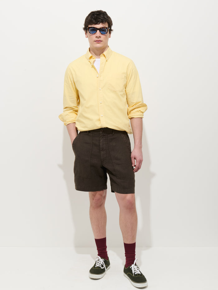 A young man wears the Mill Shirt In Cotton Poplin (yellow), dark shorts, maroon socks, green sneakers, and blue sunglasses against a white background. His hands are in his pockets as he looks forward.