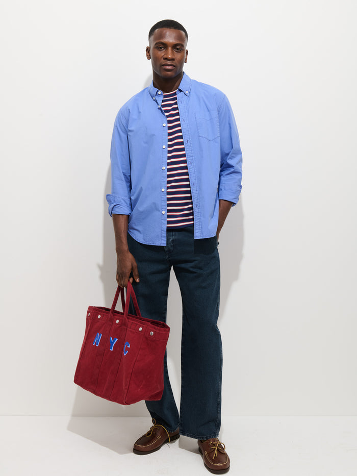 A man stands against a plain white wall wearing the Mill Shirt In Cotton Poplin—a light blue 100% cotton button-down—over a red-and-white striped tee, dark blue jeans, brown shoes, and holding a large red NYC tote bag.