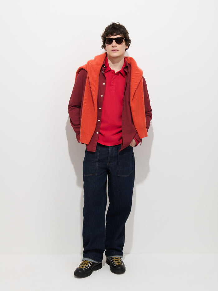Wearing sunglasses, a red polo, and the maroon Mill Shirt In Cotton Poplin—paired with dark jeans and black shoes—the individual has an orange sweater draped over their shoulders. They stand against a plain white background with hands in their pockets.