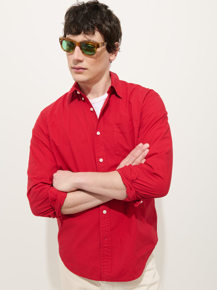 A man with dark hair, sporting green sunglasses and light pants, stands with arms crossed against a plain white background, wearing the Mill Shirt In Cotton Poplin (bright red) over a white T-shirt.