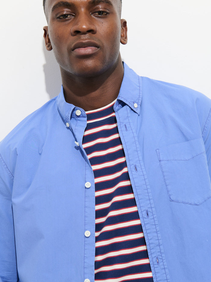 A man wears the Mill Shirt In Cotton Poplin—a 100% cotton light blue button-down—over a navy, white, and red striped tee, standing against a white background and looking at the camera with a neutral expression.