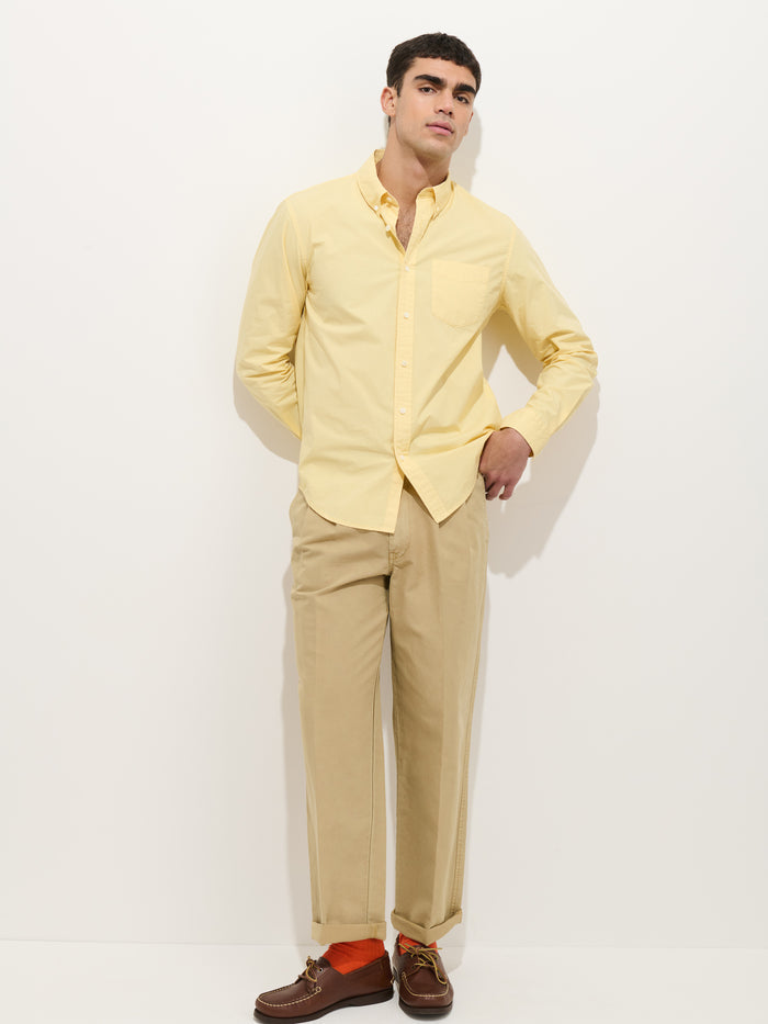 A man with short dark hair stands in front of a plain white background wearing the Mill Shirt In Cotton Poplin, light yellow, styled with beige pants, orange socks, brown boat shoes, and one hand casually in his pocket.
