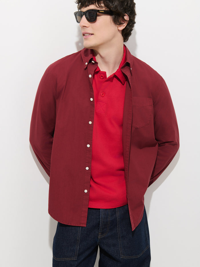 Wearing sunglasses and the Mill Shirt In Cotton Poplin—styled as a red button-up over a red polo—along with dark blue jeans, a person stands against a white background, looking to the side with their hands behind their back.