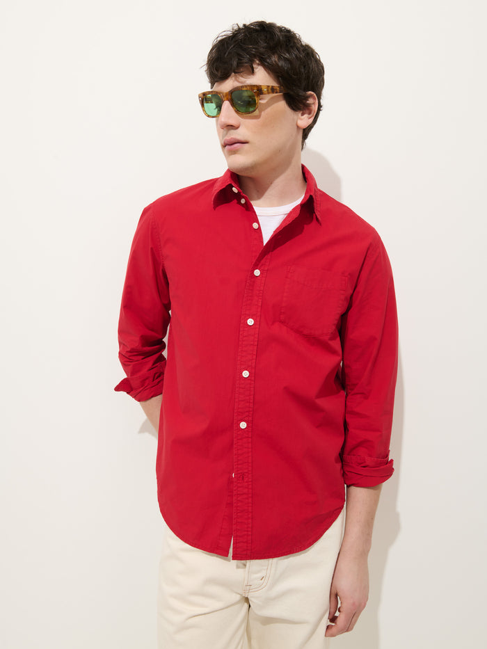 Wearing the Mill Shirt In Cotton Poplin— a vibrant red men's button-down—over a white tee, with light pants and green sunglasses, a person stands against a plain white background, one hand tucked in their pocket.