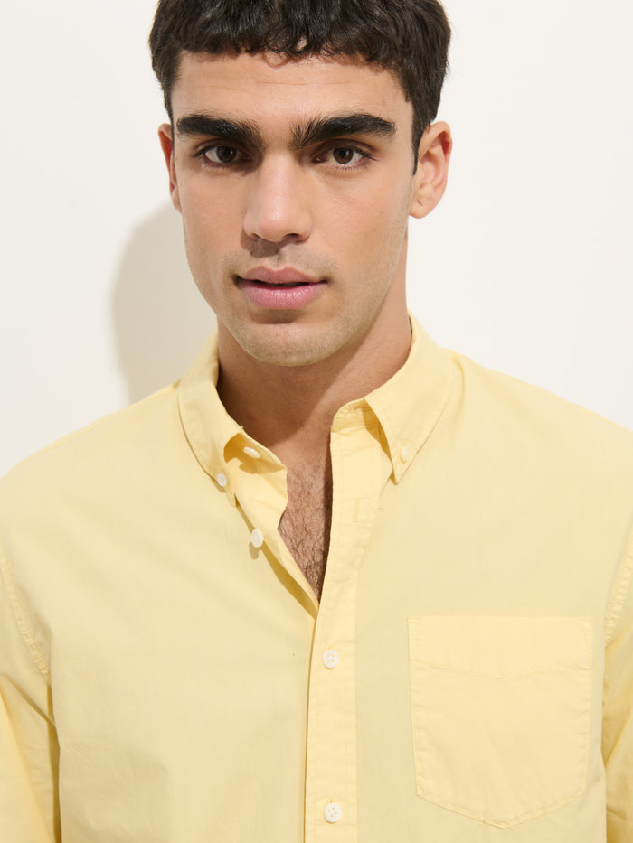 A young man with short dark hair wears the Mill Shirt In Cotton Poplin, a pale yellow 100% cotton button-down, standing against a plain white background and looking directly at the camera with a neutral expression.