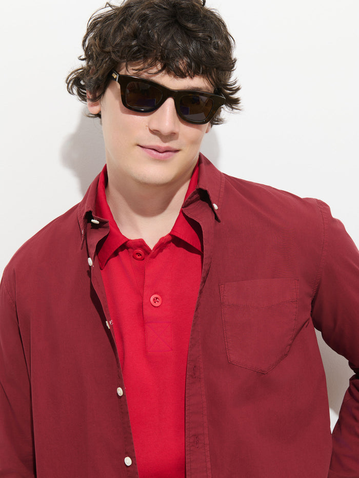 A person with wavy brown hair wears sunglasses, the Mill Shirt In Cotton Poplin in red, layered under a maroon overshirt, standing against a white background.