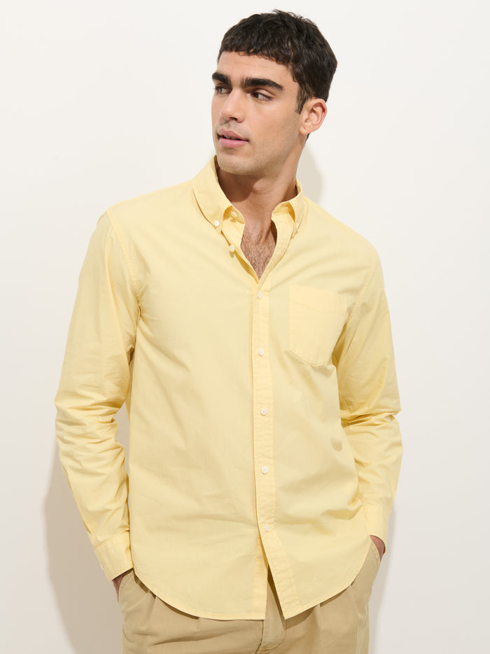 A young man with short dark hair wears the Mill Shirt In Cotton Poplin, a light yellow 100% cotton button-down, and beige pants. He stands against a white background, looking slightly left with his hands in his pockets.