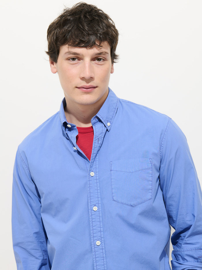 A young man with short brown hair wears the Mill Shirt In Cotton Poplin over a red t-shirt, standing against a plain white background and looking directly at the camera with a neutral expression.