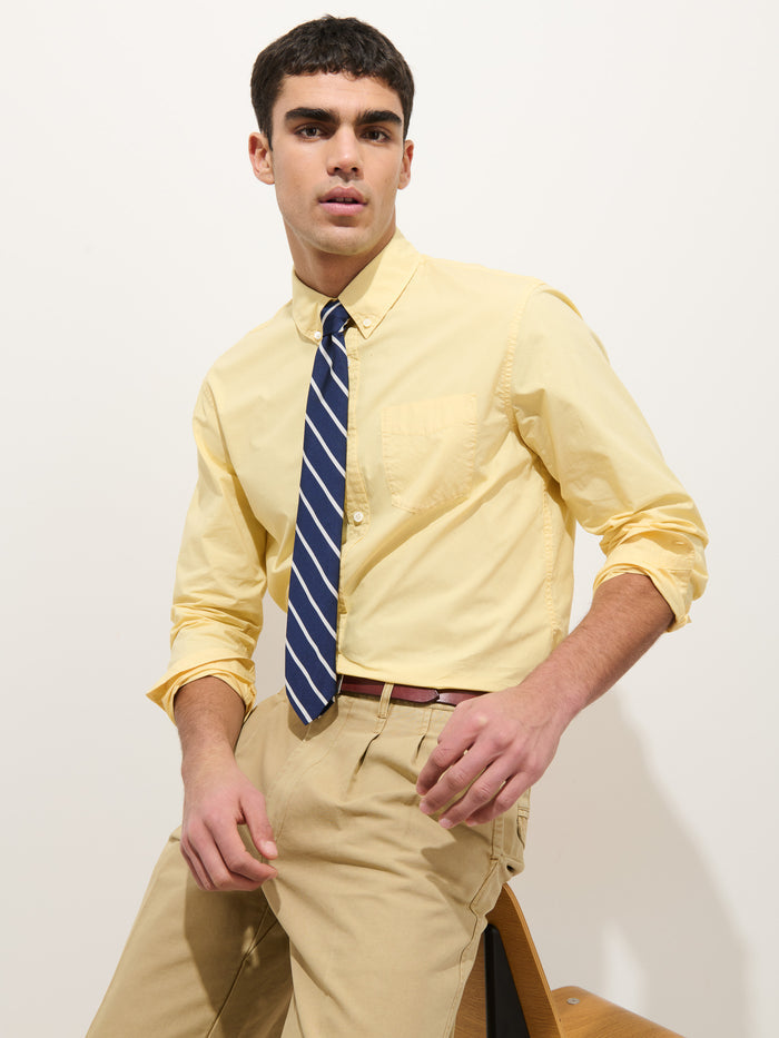 A man wearing the Mill Shirt in Cotton Poplin, a blue and white striped tie, and khaki pants sits on a wooden chair against a plain white background.