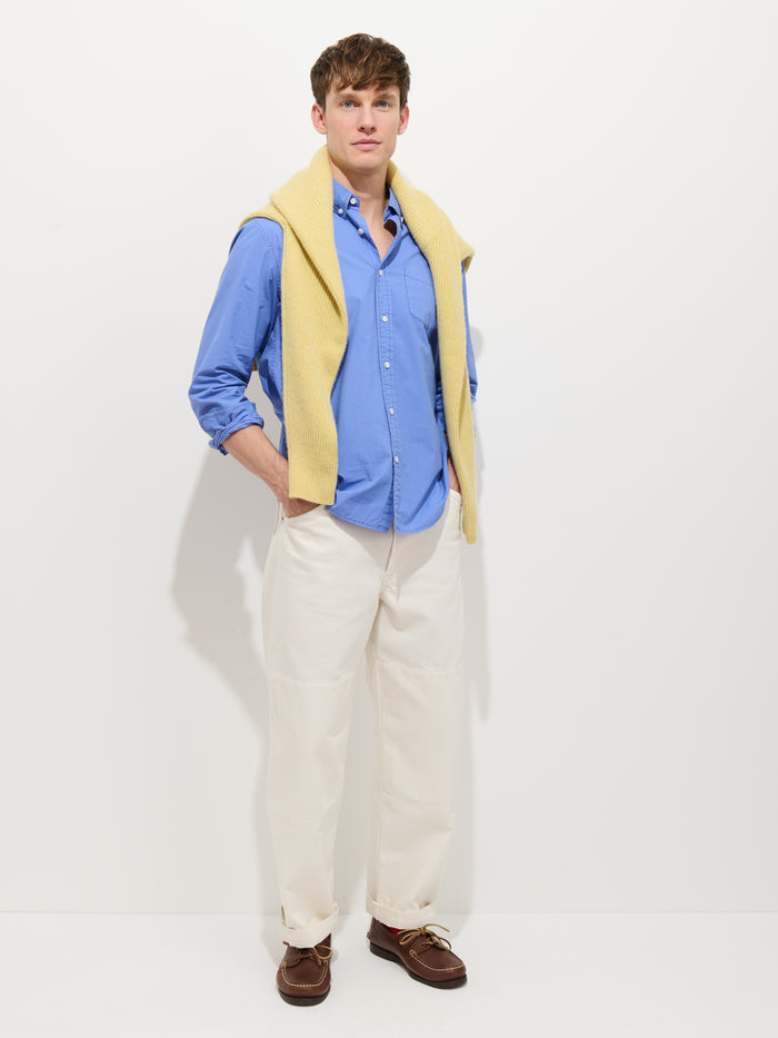 A man stands against a white background, wearing the Mill Shirt In Cotton Poplin, cream pants, brown shoes, and a yellow sweater draped over his shoulders. His hands are in his pockets as he looks forward.