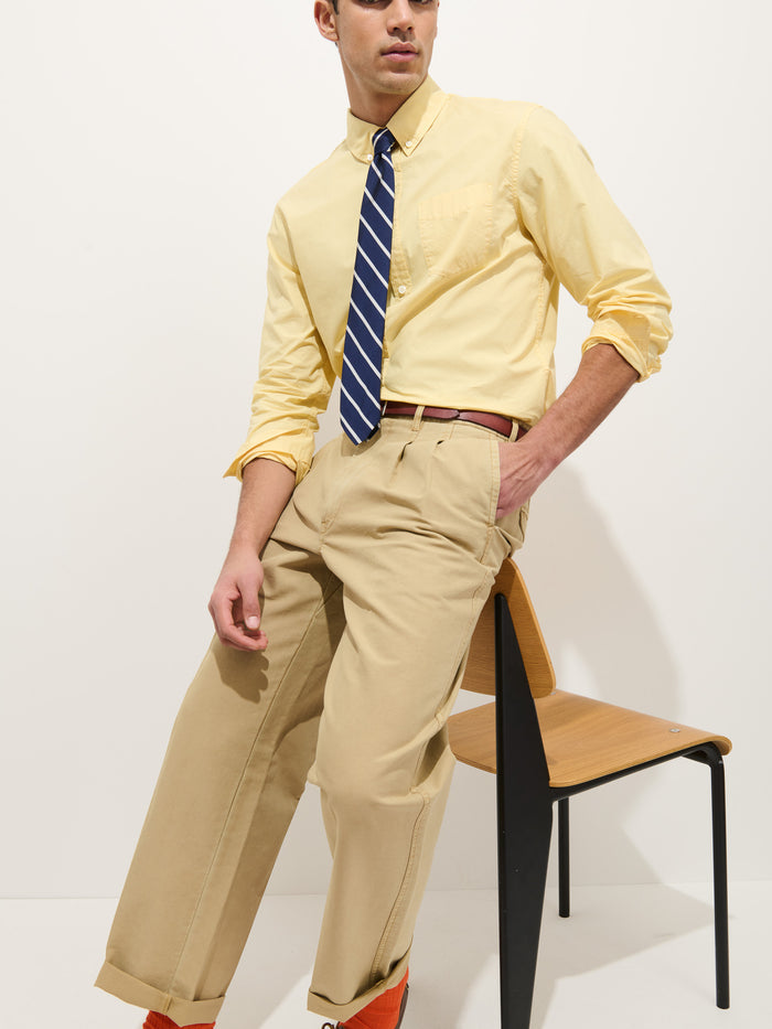 A man wearing the Mill Shirt In Cotton Poplin, a striped blue tie, beige trousers, and orange shoes sits on a wooden chair against a plain white background.