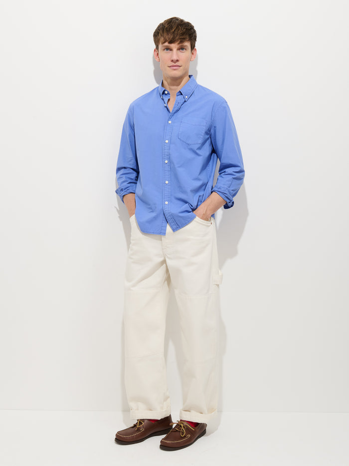 A man with short brown hair stands against a white background, wearing the Mill Shirt In Cotton Poplin with rolled-up sleeves, white wide-leg pants, and brown boat shoes. He has his hands in his pockets.