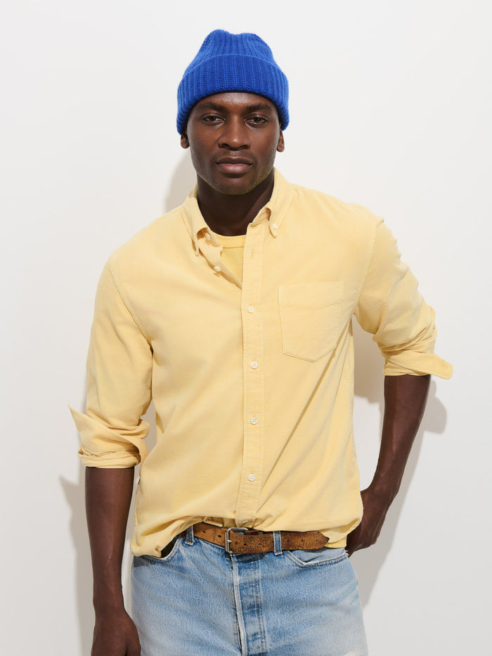 A man stands against a white background wearing a blue knit beanie, the Mill Shirt in Fine Wale Corduroy in yellow with rolled-up sleeves, a tan belt, and light blue jeans.