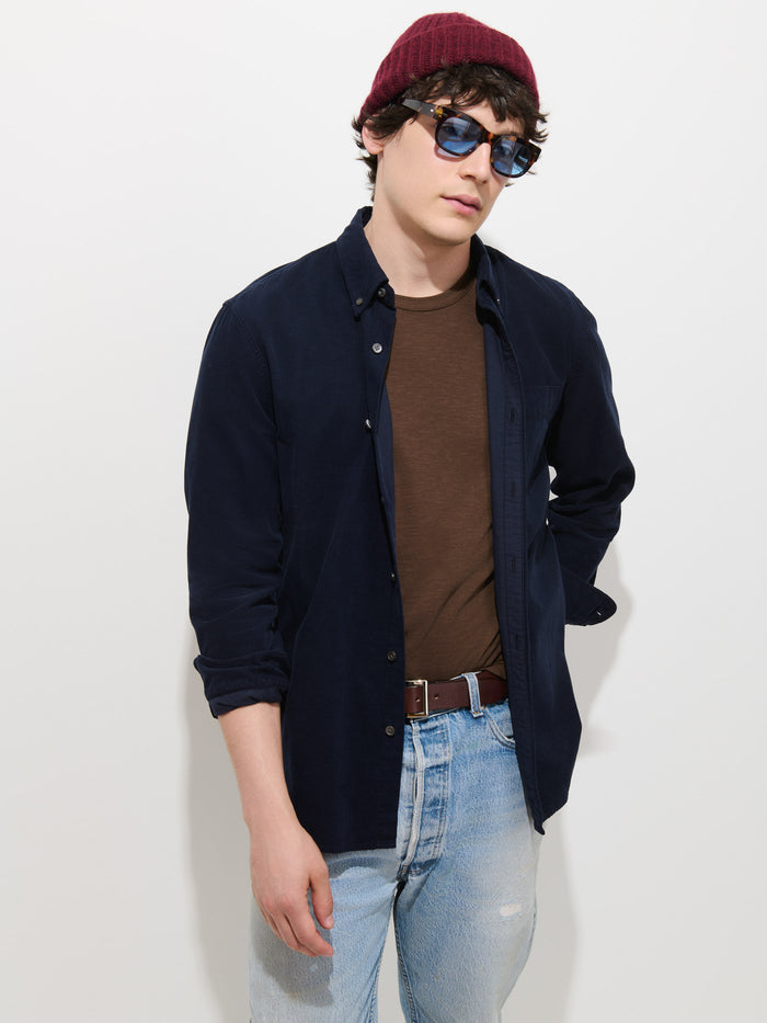 A person wearing a maroon beanie, blue sunglasses, and the Mill Shirt in Fine Wale Corduroy—crafted from 100% cotton—over a brown t-shirt and light blue jeans stands against a plain white background.
