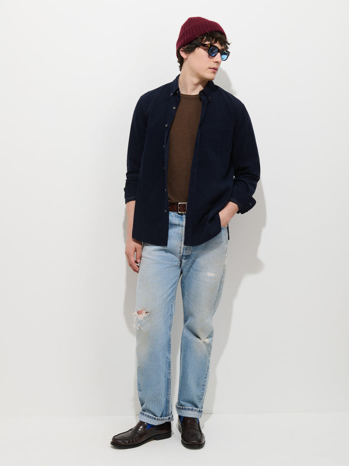 A man wearing a red beanie, sunglasses, and the Mill Shirt in Fine Wale Corduroy—styled over a brown t-shirt and paired with ripped light blue jeans and brown shoes—stands against a plain white background with one hand in his pocket.