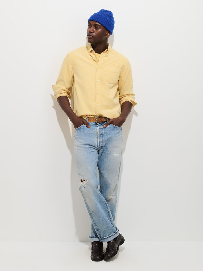 A person wearing the Mill Shirt in Fine Wale Corduroy—a yellow button-down made from 100% cotton—paired with a blue beanie, light blue ripped jeans, and black shoes stands against a white background with hands in pockets, looking to the side.