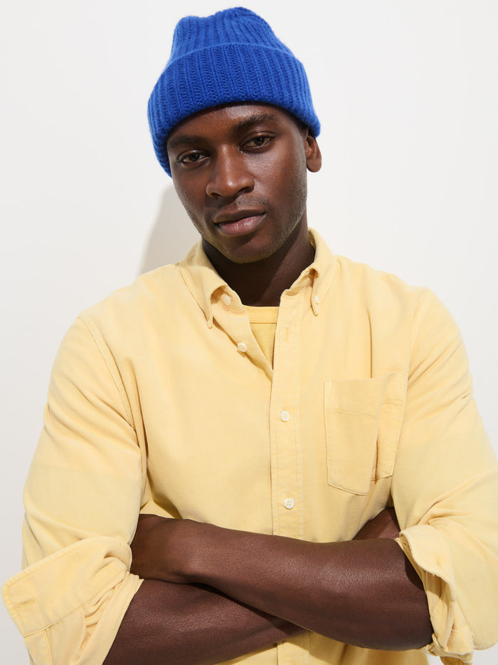 A person wearing the Mill Shirt in Fine Wale Corduroy in bright yellow and a blue knit beanie stands against a white background with arms crossed, looking at the camera with a neutral expression—the perfect shirt for effortless style.