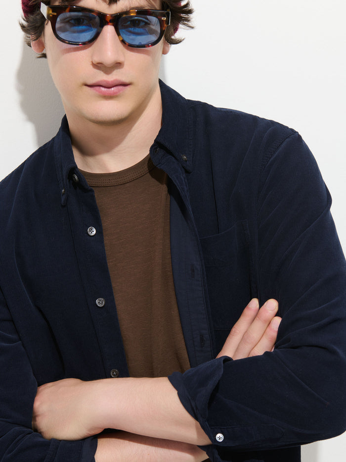 A person wearing blue sunglasses, the Mill Shirt in Fine Wale Corduroy— a dark navy men’s button-down made from 100% cotton— and a brown t-shirt stands against a white background with arms crossed, looking slightly to the side.