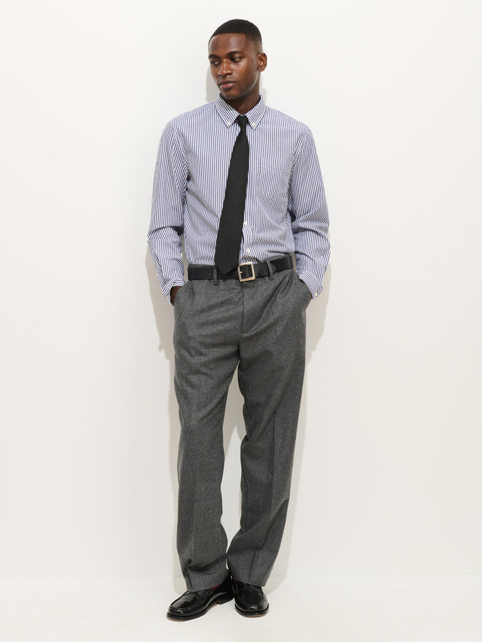 A man stands against a white background, wearing the Mill Shirt In Stripe Portuguese Poplin with a black tie, gray dress pants, and black shoes. His hands are in his pockets as he looks slightly to the side.