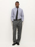 A man stands against a white background, wearing the Mill Shirt In Stripe Portuguese Poplin with a black tie, gray dress pants, and black shoes. His hands are in his pockets as he looks slightly to the side.
