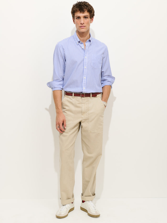 A man stands against a white background wearing the Mill Shirt in Stripe Portuguese Poplin, paired with beige pants, a brown belt, and white sneakers. One hand is in his pocket as he looks straight ahead.