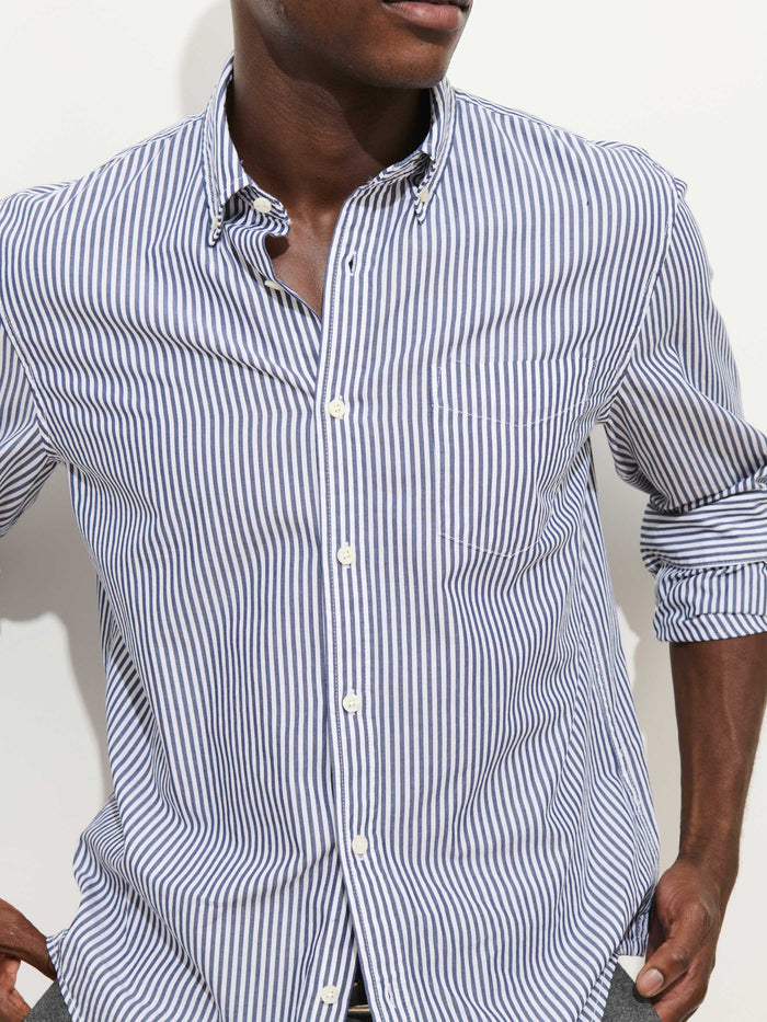 Wearing the Mill Shirt in Stripe Portuguese Poplin with blue and white vertical stripes, a person stands against a plain light background, sleeves rolled up and face partially out of frame.