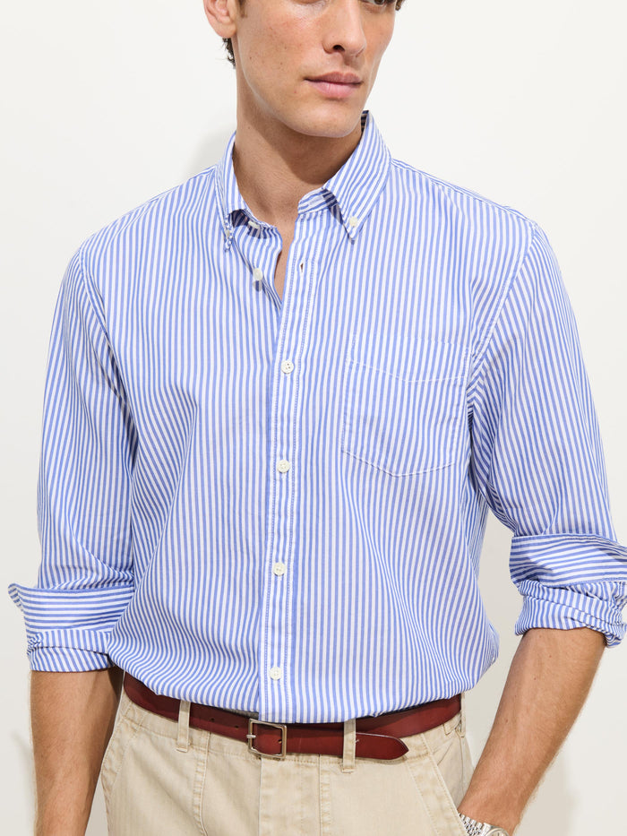 A man wears the Mill Shirt in Stripe Portuguese Poplin—with blue and white vertical stripes, lightweight fabric, and rolled-up sleeves—paired with beige pants and a brown belt, standing against a plain light background.