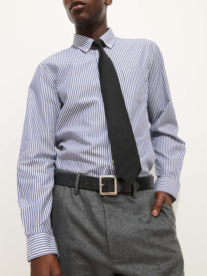 A person stands against a plain light background with one hand in their pocket, wearing the Mill Shirt In Stripe Portuguese Poplin, paired with a black tie, grey pants, and a black belt with a silver buckle.