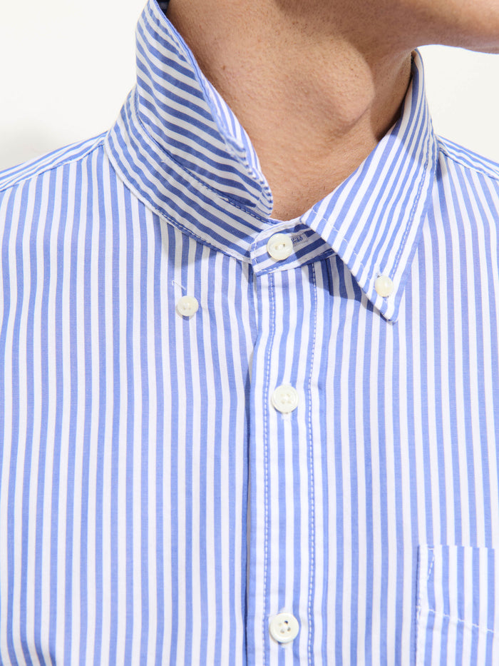 A close-up of a person wearing the Mill Shirt In Stripe Portuguese Poplin, featuring crisp blue and white vertical stripes with a buttoned collar, showing part of the neck and jawline.