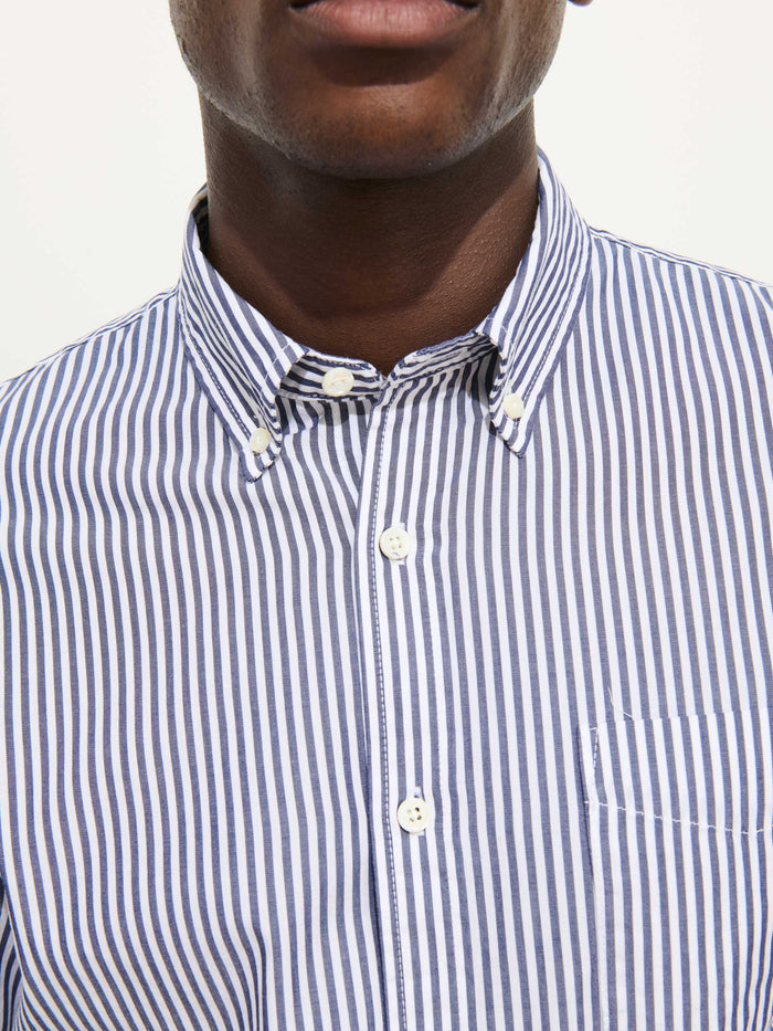 A person wearing the Mill Shirt in Stripe Portuguese Poplin—a men's cotton shirt with blue and white vertical stripes and a button-down collar. The image is cropped to show the lower half of the face and upper torso.
