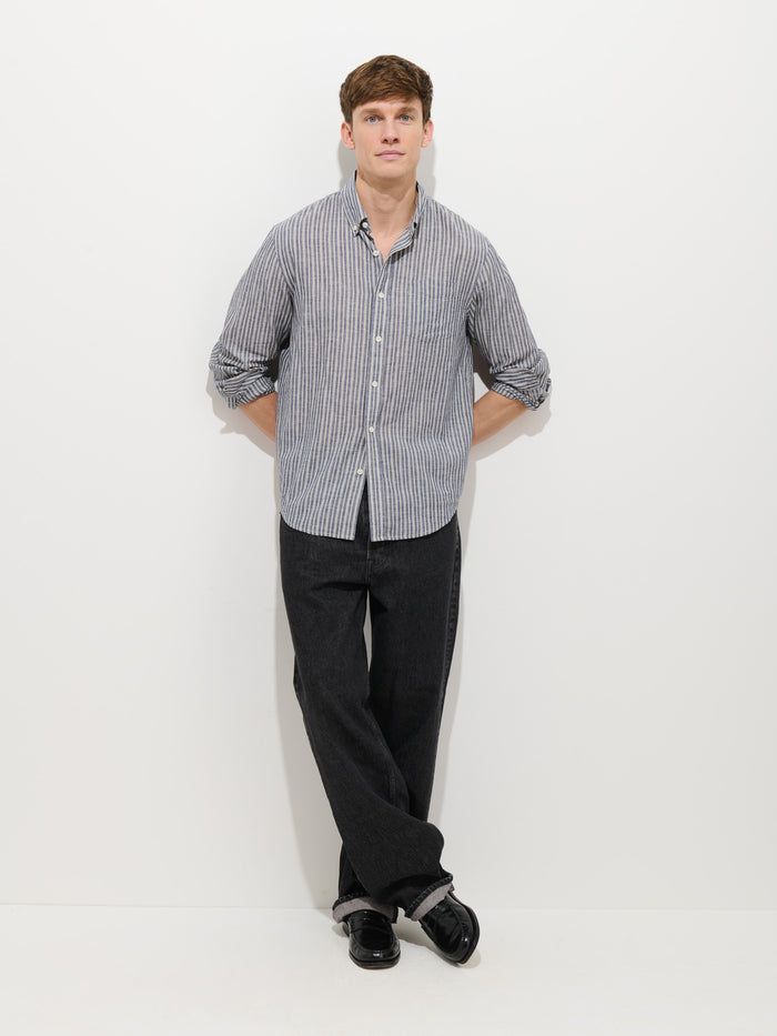 A man stands against a white wall with his hands behind his back, wearing the perfect shirt—the Mill Striped Shirt In Chambray—paired with loose black pants and black shoes.