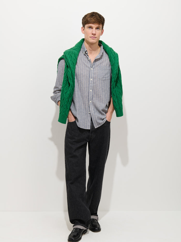 A man stands against a white background wearing the Mill Striped Shirt In Chambray, a green sweater draped over his shoulders, loose black pants, and black shoes, with his hands in his pockets.