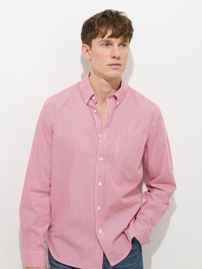 A young man stands against a white background, wearing the Mill Striped Shirt in cotton poplin—red and white stripes—and blue jeans, with one hand in his pocket and a neutral expression.
