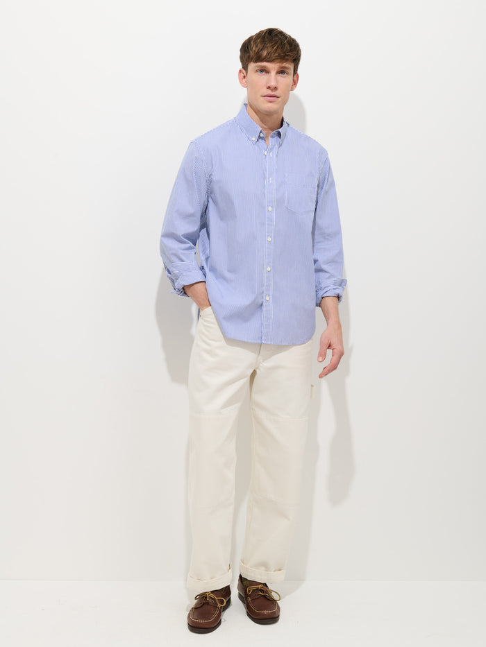 A man stands against a plain white background wearing the Mill Striped Shirt In Cotton Poplin, paired with off-white pants and brown boat shoes, with one hand in his pocket.
