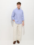 A man stands against a plain white background wearing the Mill Striped Shirt In Cotton Poplin, paired with off-white pants and brown boat shoes, with one hand in his pocket.