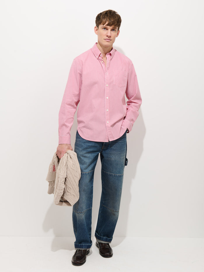 A young man stands against a white background, wearing the Mill Striped Shirt in Cotton Poplin, blue jeans with visible seams, and dark shoes. He holds a light-colored cable-knit sweater in his left hand.