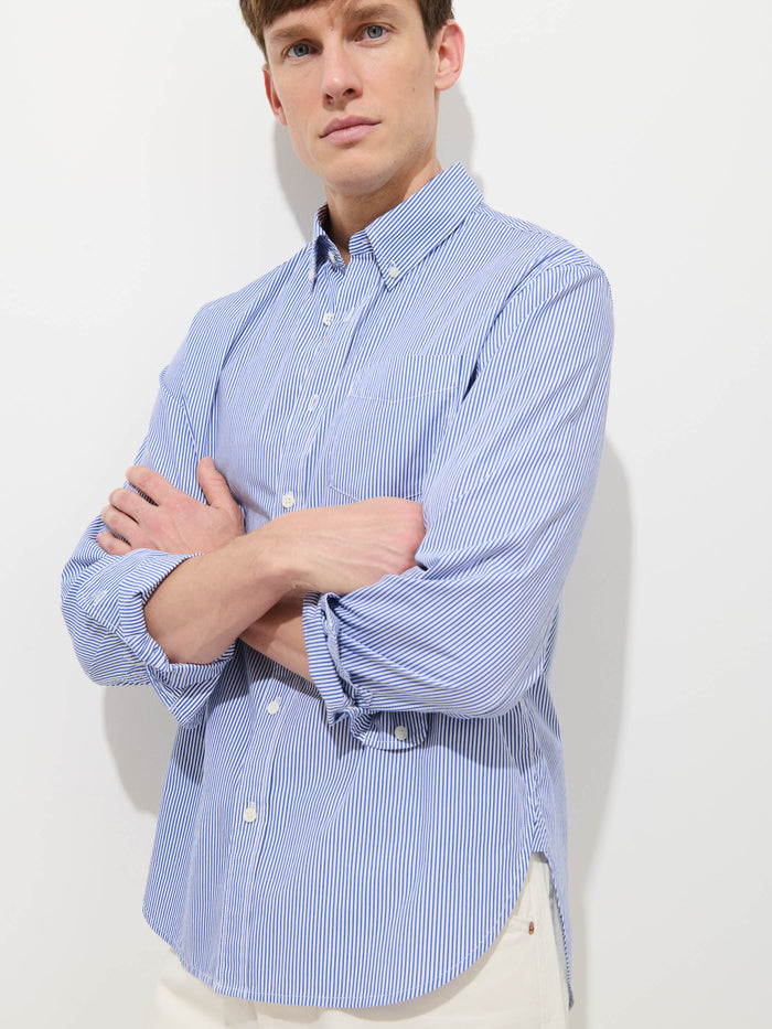 A man wears the Mill Striped Shirt in Cotton Poplin, featuring blue and white stripes, standing against a plain white background with his arms crossed and a neutral expression.