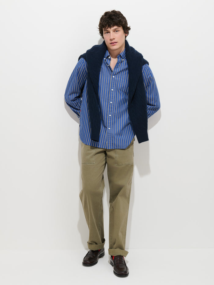 A young man stands against a white wall wearing the Mill Striped Shirt in Cotton Poplin, paired with khaki pants, brown shoes, and a navy sweater draped over his shoulders.