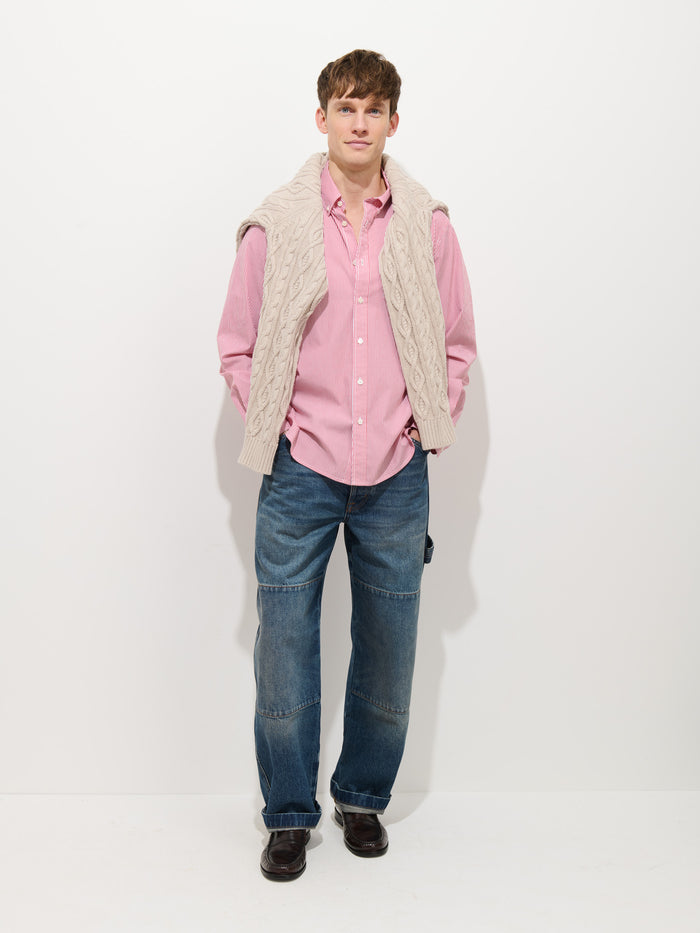 A man stands against a plain white background, wearing the Mill Striped Shirt in Cotton Poplin, blue jeans, dark shoes, and a cream-colored cable knit sweater draped over his shoulders.