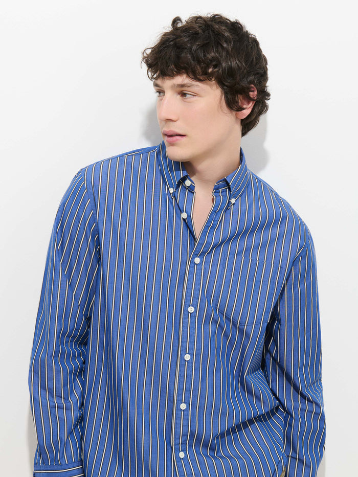 A young person with short, curly brown hair wears the Mill Striped Shirt in Cotton Poplin—featuring perfect blue fabric with white and yellow vertical stripes—as they look to the side against a plain white background.