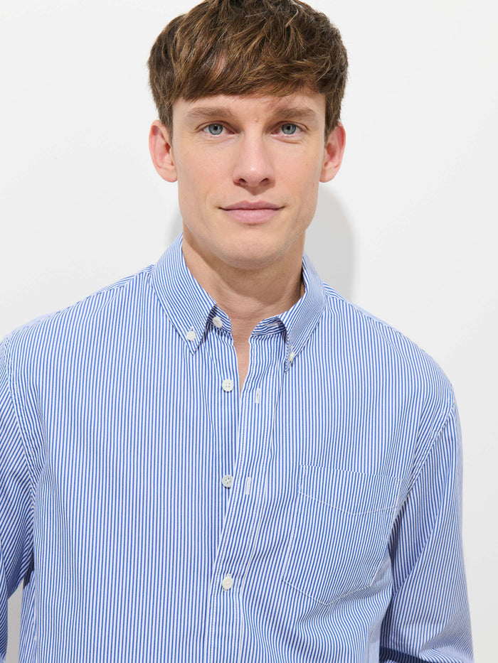 A man with short brown hair wears the Mill Striped Shirt in Cotton Poplin, a light blue and white 100% cotton button-down, standing against a plain white background and looking directly at the camera with a neutral expression.