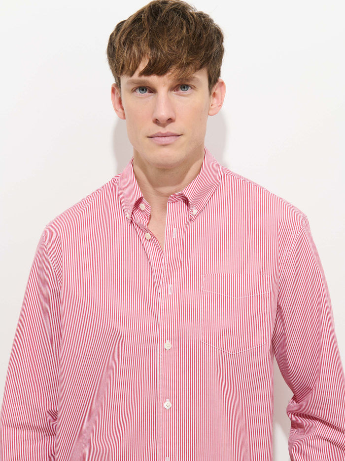 A young man with short brown hair and blue eyes wears the Mill Striped Shirt in Cotton Poplin—a red and white vertically striped 100% cotton button-down—standing against a plain white background, looking directly at the camera with a neutral expression.