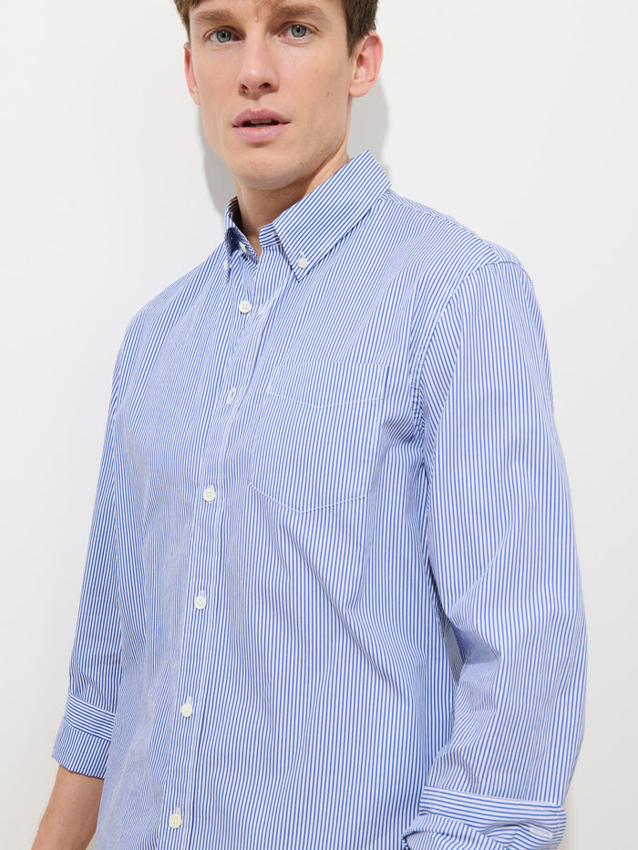 A man wears the Mill Striped Shirt in Cotton Poplin, featuring blue and white stripes. He stands against a plain white background, looking slightly to the side—this versatile shirt is perfect for any occasion.