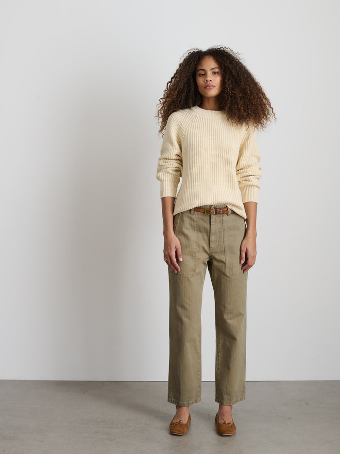 A person with curly hair stands in front of a white wall, wearing The Neil Pant In Herringbone, a cream-colored cotton sweater, brown belt, and brown shoes. They have a neutral expression and their arms are at their sides.